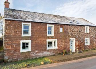 Stone built front exterior of a traditional Cumbrian cottage overlooking a quiet road.