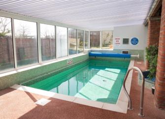 An indoor heated pool with a surrounding terrace. Floor-to-ceiling windows line one side.