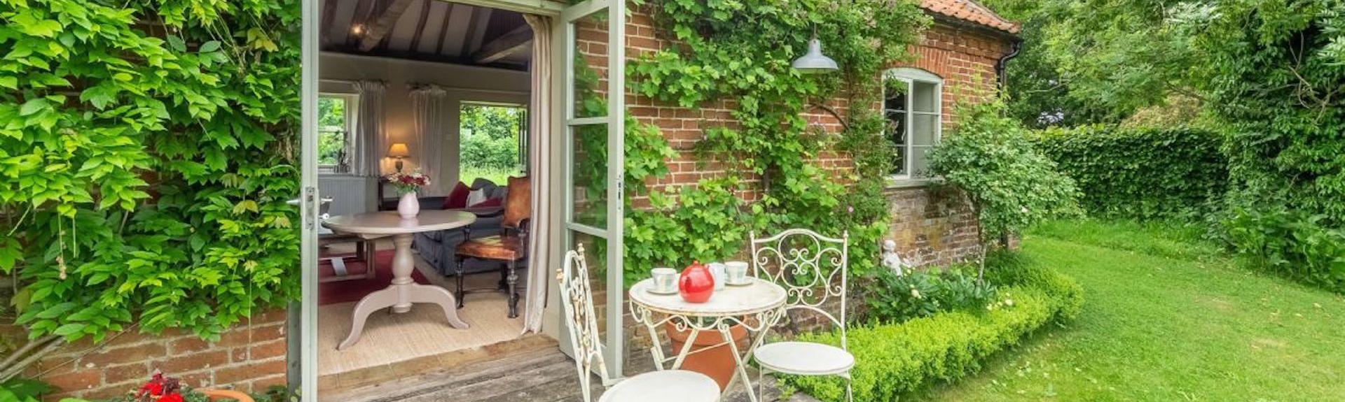 A single storey cottage with wysteria creeping around open French windows. Outside is a deck with tables and chairs overlooking a lawned garden..