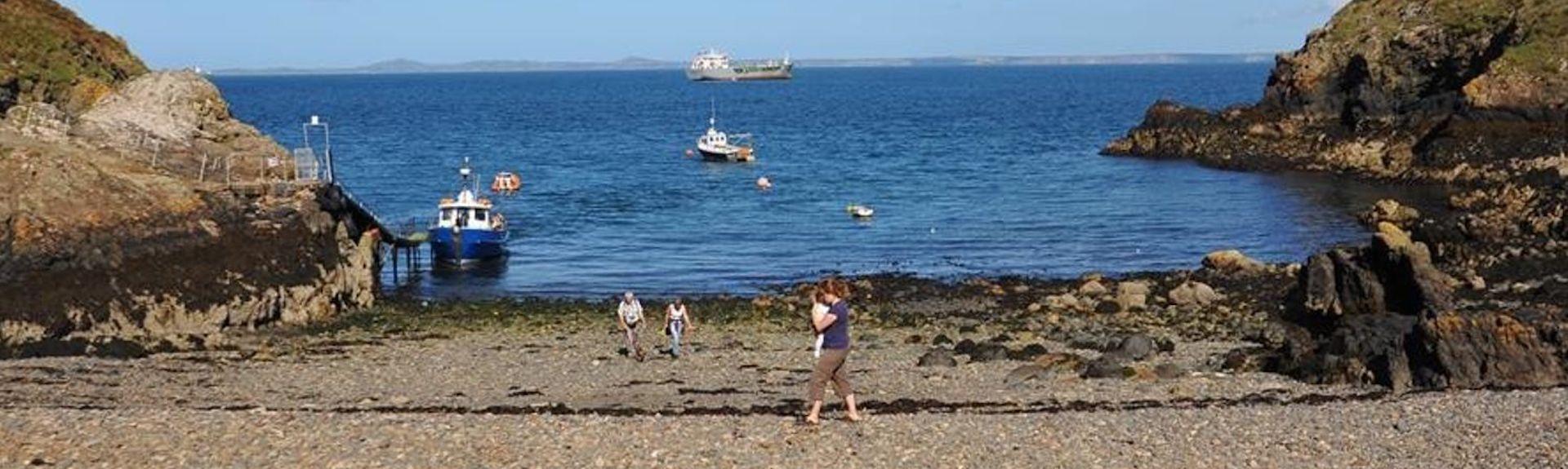 A shingle beach with an upturned rowing boat in the foreground and people walking nestles between two rocky headlands. A small fishing boat is moored to a jetty. Out to sea a large tanker heads towards the horizon.
