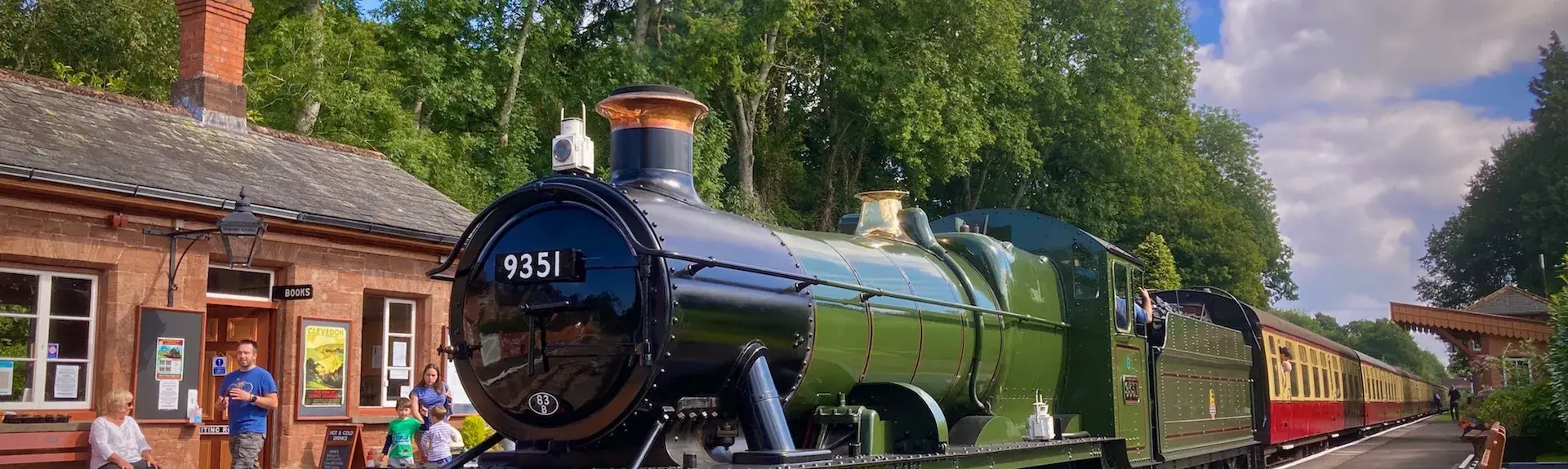 A steam engine waits to depart with its train on a sunny day in Crowcombe 