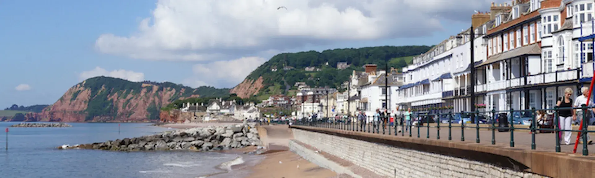 Sidmouth's seafront with the ocean, a sloping pebble beach backed by a long esplanade. Behind this is a row of Georgian hotels and cafes.