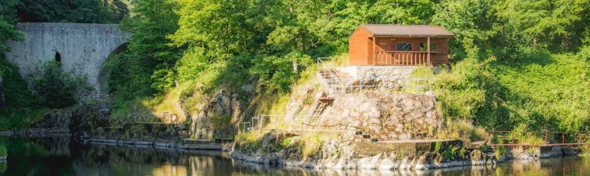 a wooden lodge in Aberdeenshire backed by trees is reflected in the calm waters of a river below it.