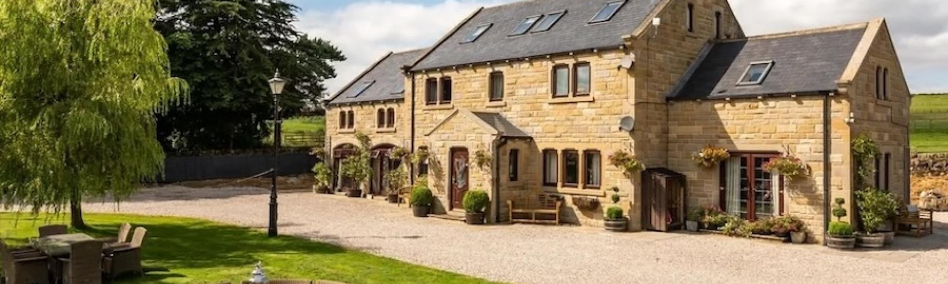 An extended stone-built farmhouse with a large drive and lawn surrounded by countryside.