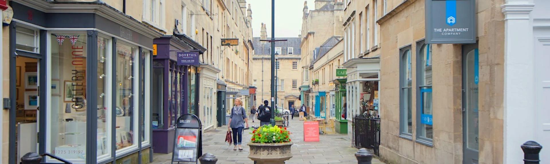 A pedestrianised Georgian street with lamp posts and floral displays down its centre bordered by trendy 3-storey houses and shops bult of Bath stone