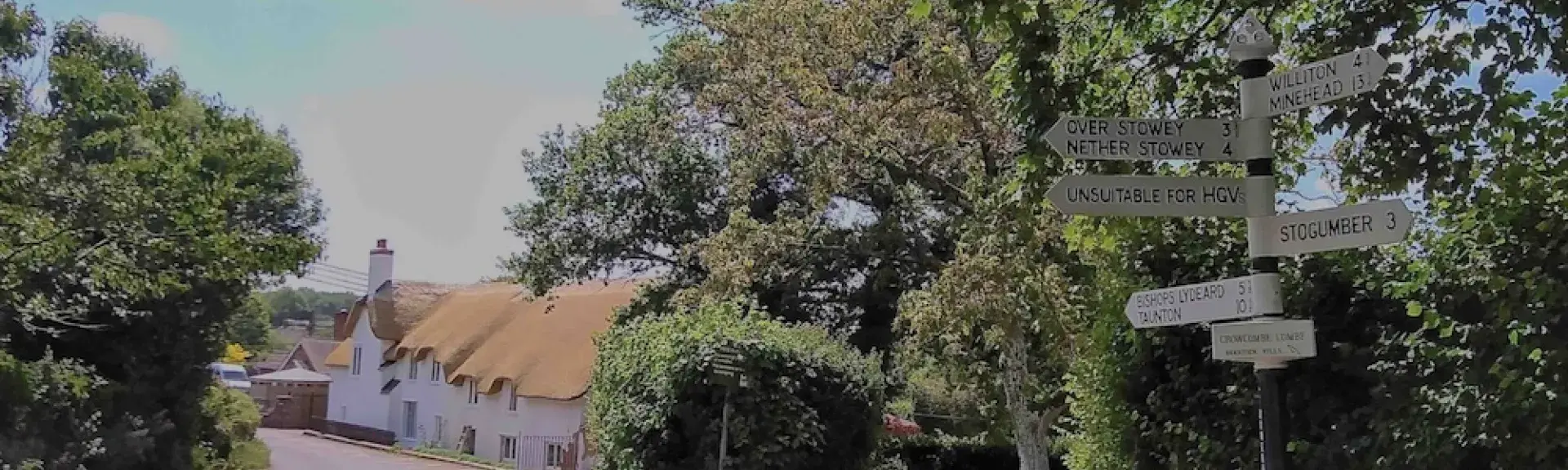 Crowcombe village street. In the foreground is a signpost with directions to local villages. Behind it are trees and a newly thatched row of cottages.