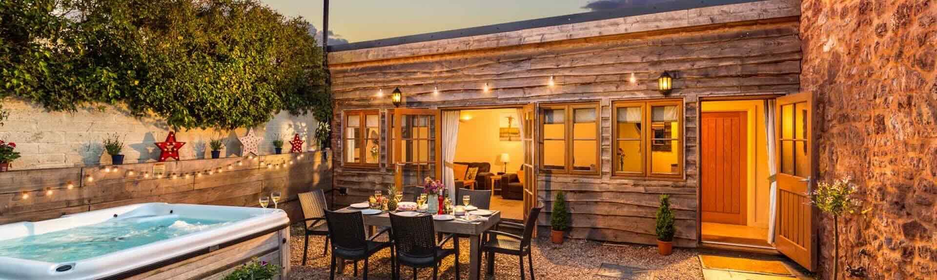 A wood-clad single-storey Somerset holiday cottage with a table laid for dinner in a courtyard at dusk.