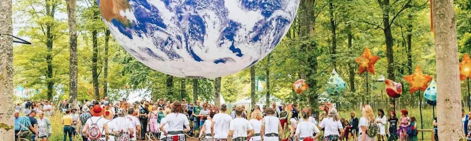 Festival goers in garish attire admire a large inflatable globe hanging from woodland trees in Kendal.