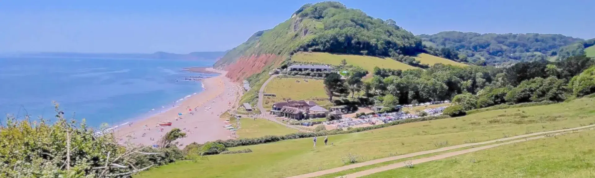 Branscombe beach in Devon. A field slopes down to a cluster of sea shanties overlooking a wide shingle beach, beyond which the lad rises to form cliffs topped by woodland. 