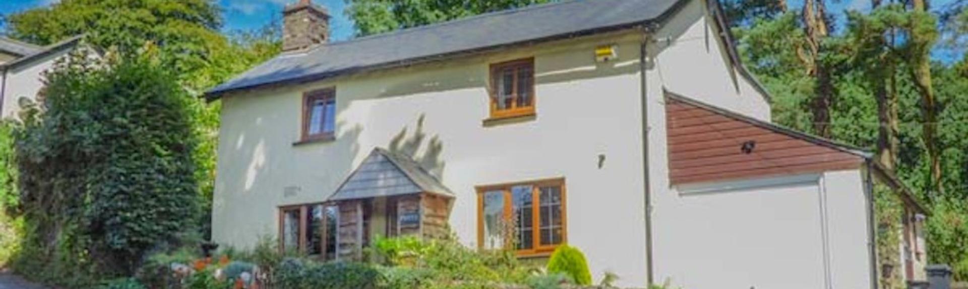 The exterior of a double-fronted Exmoor holiday cottage overlooks a front garden and is surrounded by trees.