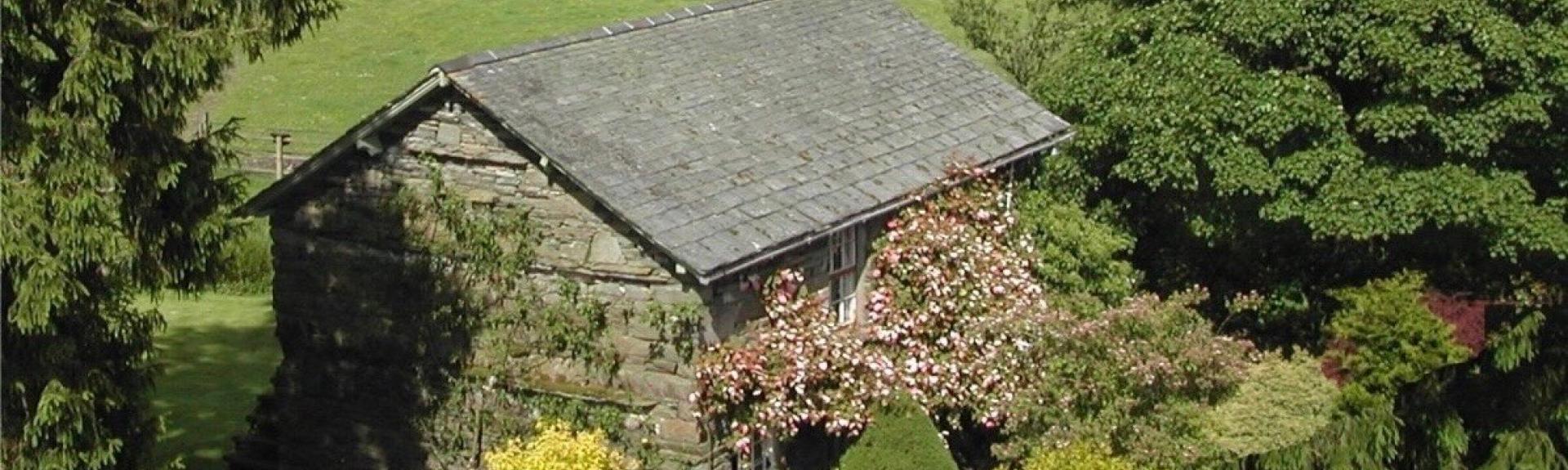 A stone-built, slate-roofed Lake District holiday cottage surrounded by shrubs and small trees with hilly fields in the distance.