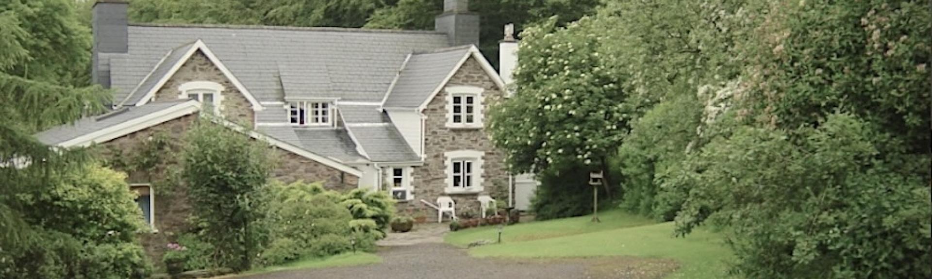 A sweeping drive approaches a twin-gabled Exmoor holiday cottage half-surrounded by mature trees.