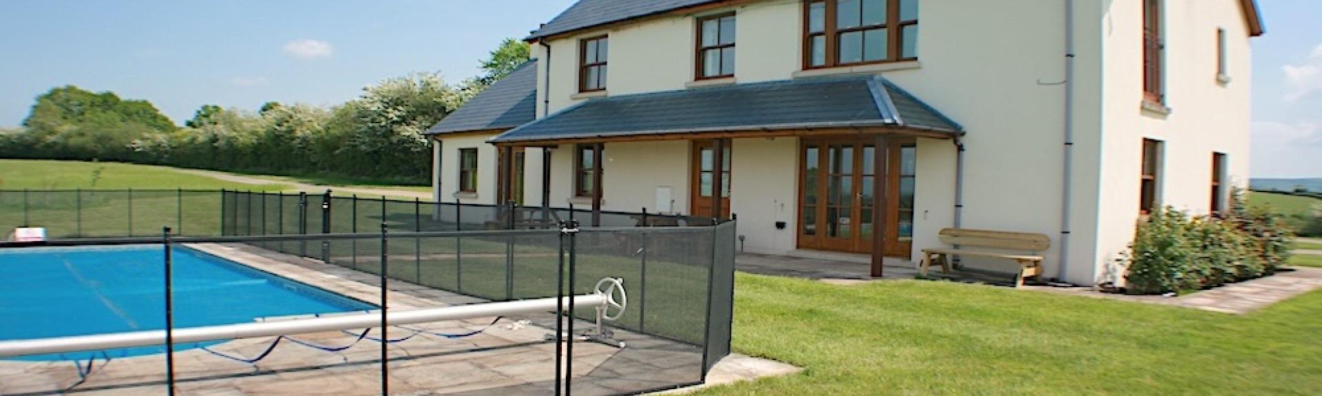 A fenced-in outdoor pool on a lawn overlooked by a large holiday cottage.