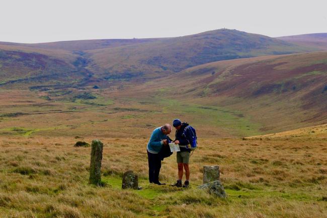 Two Dartmoor walkers study maps next to a standing stone in an open expanse of of moorland with a large hilly, moorland Tor in the background