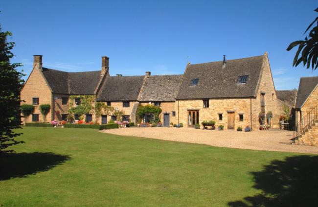 An old Manor House in the Cotswolds with honey-colours bricks and high slate roof overlooks a shingled drive and large lawn.s