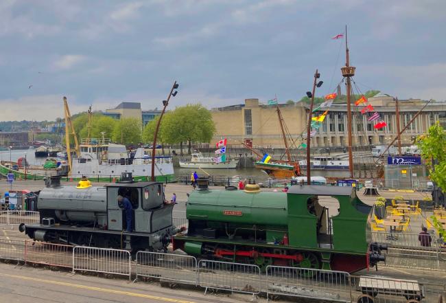 Two saddle tank steam engines simmer quietly on a Bristol Quayside. In the background are a variety. of boats and sailing ships moored to the quayside.