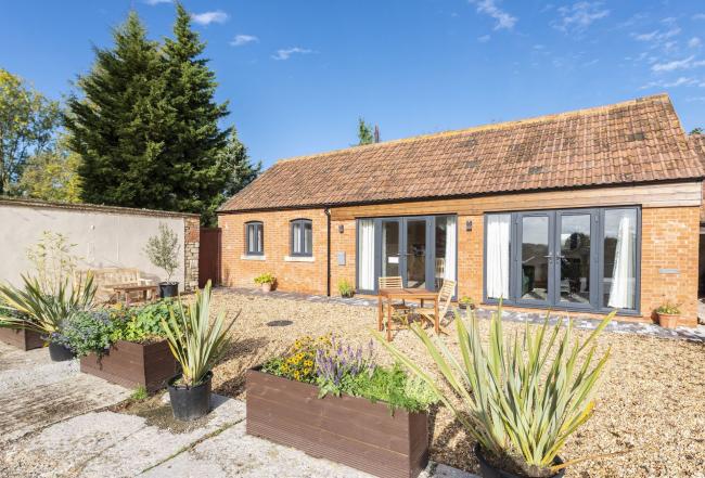 A single storey holiday home with French windows overlooks a large courtyard with outdoor seating and tubs of flowers.