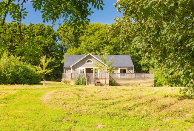 The exerior of a wooden holiday lodge and deck against a sunlit back drop of trees overlooking a field.