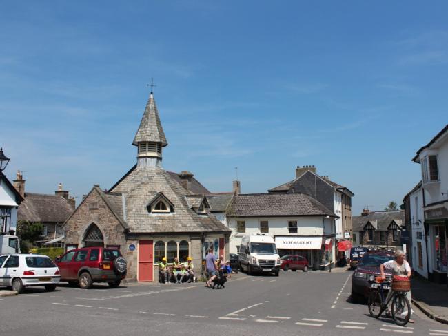 An old circular mrket building with a conical tower surrounded by small shops and parked cars in Chagrod, Devon.