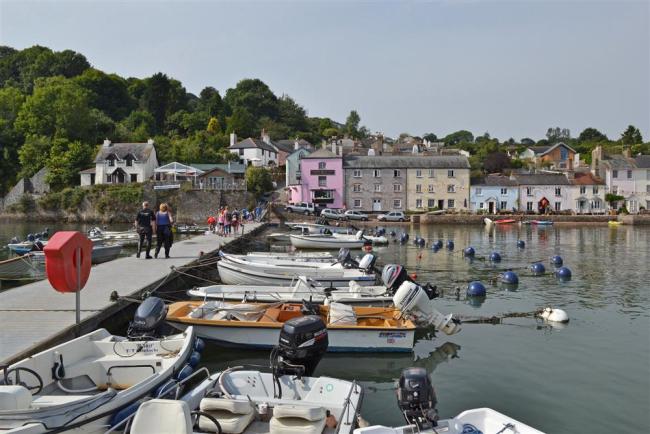 A floating pier extends into the River Dart Estuary. Smll boats are moored against the pier. Pastel coloured buildings line the quayside in the background