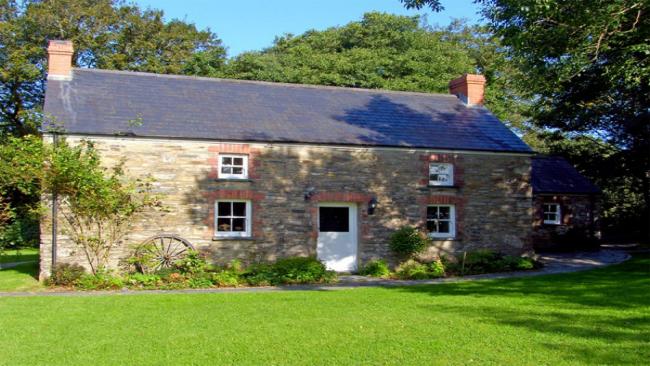 The exterior of  stone-built Welsh farmhouse overlooking  lawn with tall tree to its side and rear. an old cart wheel leans against the wall of the house.