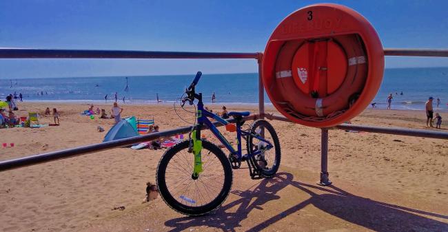 A bicycle is tethered to a railing alongside a life buoy overlooking a large sandy beach on which people are sunbathing. 