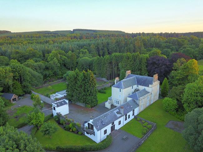 aerial view of a large country house with attached outbuuildings surrounded by lawns and woodland