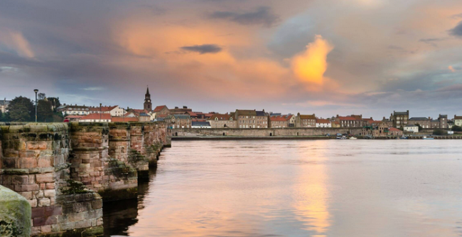 A waterside view at sunset in Berwick-upon-Tweed. The stone buttresses of anold bridge lead towards the far bank lined with old 3-storey houses.