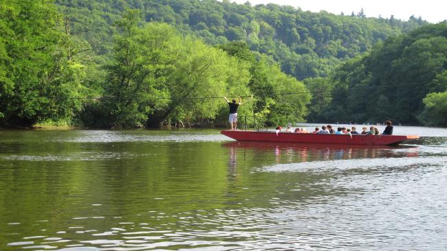 A long punt operating as a ferry crosses the wide River Wye surrounded by tree-lined banks