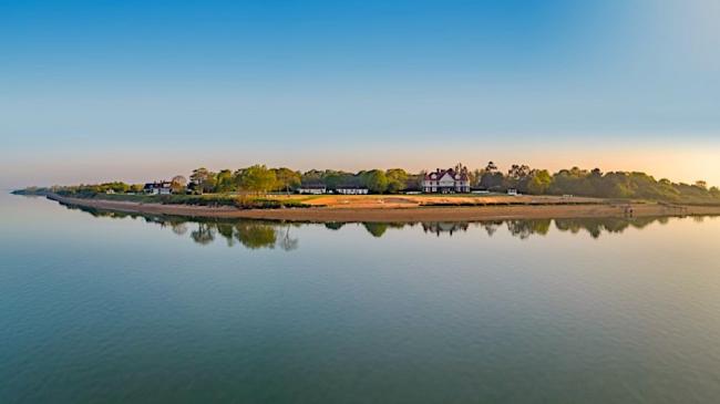 A low Island with trees. A handful of Essex holiday cottages and sandy beaches are reflected in a calm sea