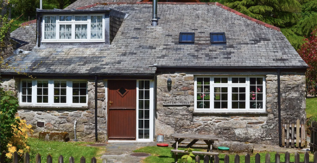 A slate roofed holiday cottage with dormer windows overlooks a lawn and picket fence.