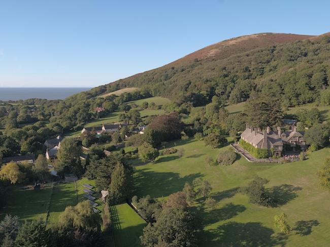 An aerial view of a small hamlet nestling beneath a tree and bracken covered moorland hill top.