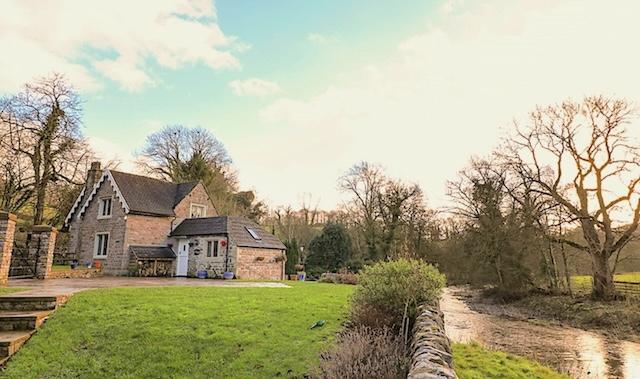 A river flows past a field and curves round a stone house in the distance.