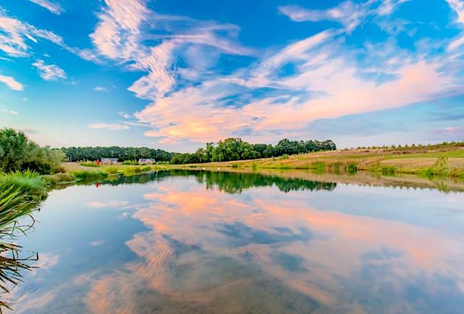 Two Piddinghoe holiday lodges backed by woods overlook s large lake in which a sunset is reflected.