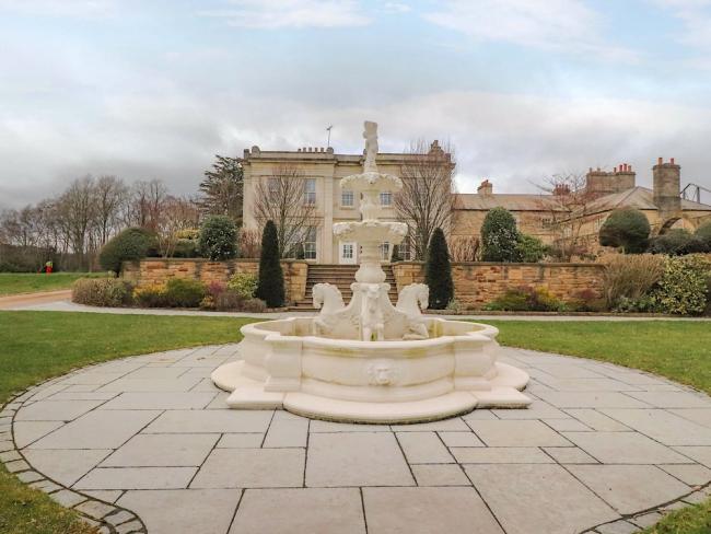 An ornamental fountain stands in front of a country house half-hidden behjind a wall.