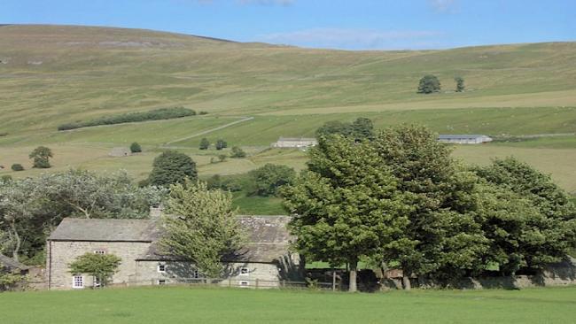 A large farmhouse partly surrounded by trees, sits beneath moorland hillside.