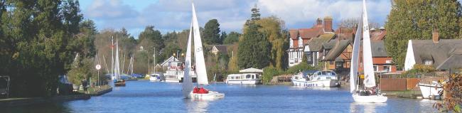 Sailing dinghies tack up a wide river with other boats moored along its banks