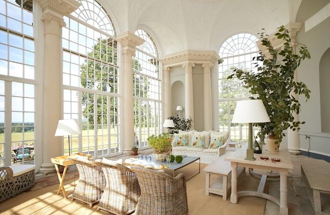 A luxuriously furnished, tall-windowed orangery with table and whicker chairs.