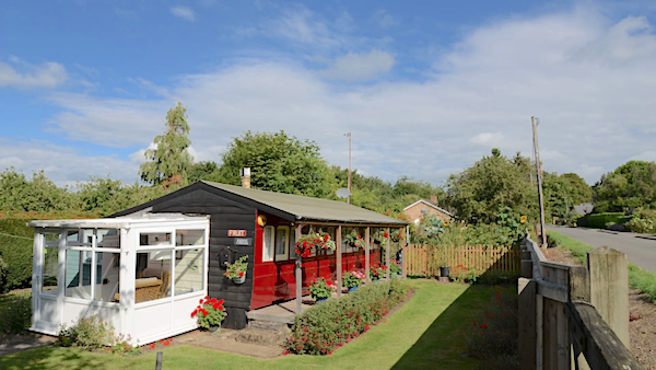 A restored old railway carriage converted to a self-catering holiday home festooned with flowers stands in a well-kept garden in Wisbech on a sunny day