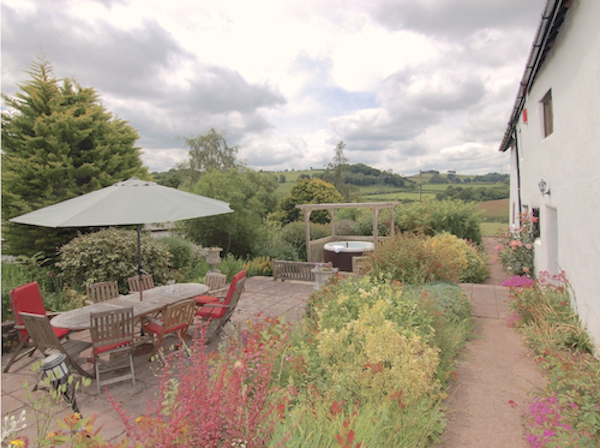 A flower-fill country garden with a parasol-covered wooden dining table and chairs.