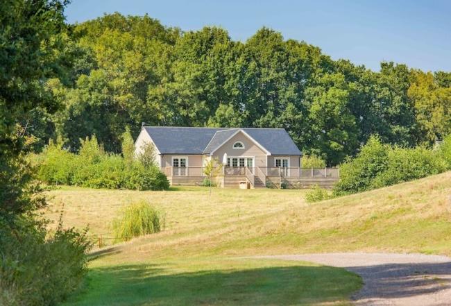 A New England style wooden holiday lodge. In the foreground are sloping fields while behind the lodge is a wood.