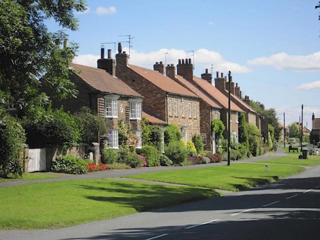 A row of traditional cottages overlook a wide grassy bank above a quiet village street in North Yorkshire