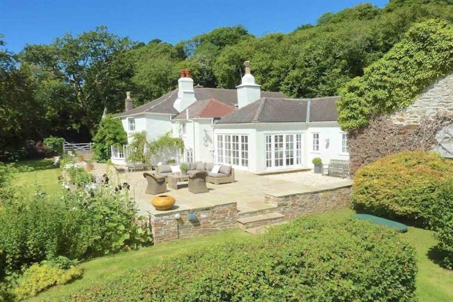 The exterior of a large holiday cottage with a single-storey extension and a spacious raised patio is surrounded by trees and shrubs.