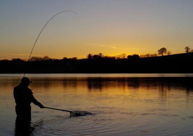 Silhouetted by a setting sun, an angler on a fishing holiday lands a fish