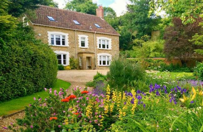 A stone-built, double-fronted country house is surrounded by trees, shrubs and flower-filled gardens.