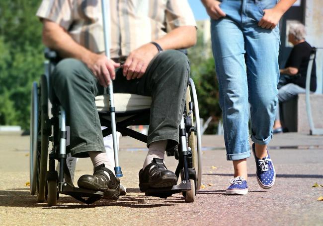 The lower half of a person sitting in a wheelchair with the lower half of a woman in jeans walking alongside.