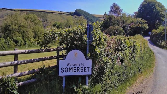 A ‘Welcome to Somerset’ road sign stands on a winding country lane that curls into the distance.