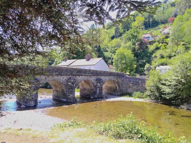 A wide, shallow river flows under an old 4-arch stone bridge.