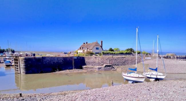 Two yachts, overlooked by thatched cottages await an incoming tide to access a small harbour.