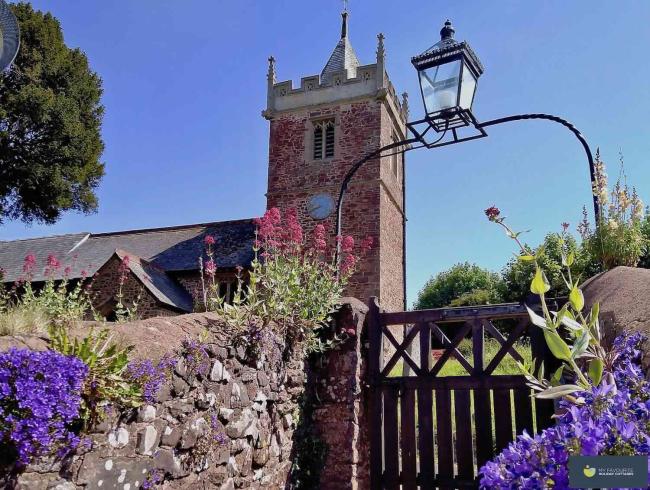 A church gate with an overhead lantern leads to a red sandstone Somerset church with a tower.